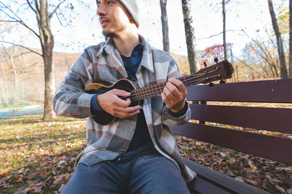 Tutku, sonbahar yaprakları arasında parktaki ahşap bir bankta oturup ukulele çalıyor. Etrafı ağaçlarla çevrili ve yumuşak güneş ışığıyla yıkanmış, yaratıcılıkla dolu huzurlu bir anın tadını çıkarıyor..