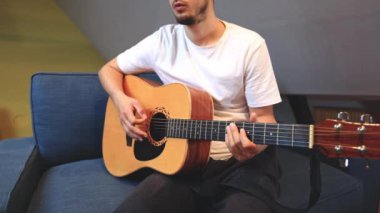 Müzisyenlerin elleri nazikçe akustik gitar tellerini tıngırdatıyor, evin içindeki sıcak ışık parlayan ahşabı yansıtırken karmaşık hareketleri ve hassasiyeti yakalıyor.