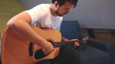 Müzisyenlerin elleri nazikçe akustik gitar tellerini tıngırdatıyor, evin içindeki sıcak ışık parlayan ahşabı yansıtırken karmaşık hareketleri ve hassasiyeti yakalıyor.