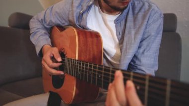 Müzisyenlerin elleri nazikçe akustik gitar tellerini tıngırdatıyor, evin içindeki sıcak ışık parlayan ahşabı yansıtırken karmaşık hareketleri ve hassasiyeti yakalıyor.
