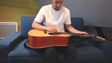 Müzisyenlerin elleri nazikçe akustik gitar tellerini tıngırdatıyor, evin içindeki sıcak ışık parlayan ahşabı yansıtırken karmaşık hareketleri ve hassasiyeti yakalıyor.