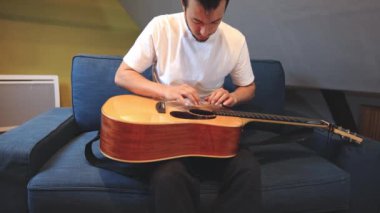 Müzisyenlerin elleri nazikçe akustik gitar tellerini tıngırdatıyor, evin içindeki sıcak ışık parlayan ahşabı yansıtırken karmaşık hareketleri ve hassasiyeti yakalıyor.