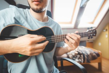 Tavan pencerelerinden geçen doğal ışık altında siyah bir ukulele çalan bir adamın yakın çekimi. Ukulele çalmanın ve kendini yaratıcı ruha daldırmanın tadını çıkarıyorum..