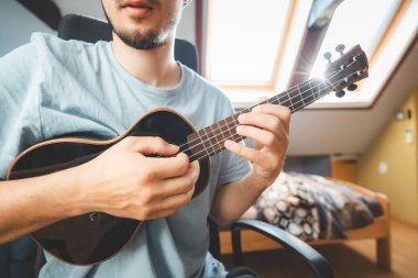 Close-up of a man playing a black ukulele under natural light streaming through skylights. Enjoying the feeling of playing the ukulele and immersing oneself in the creative spirit. clipart