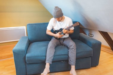Yetenekli bir adam mavi bir kanepede oturur, dikkatlice siyah bir ukulele ayarlar. Müzisyen, ukulele çalmanın ve kendini yaratıcı ruha daldırmanın tadını çıkarıyor..