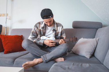 Man with talent sits on a gray sofa, carefully tuning a black ukulele. Musician is enjoying the feeling of playing the ukulele and immersing oneself in the creative spirit. clipart