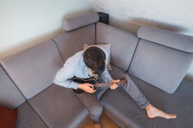 Man with talent sits on a gray sofa, carefully tuning a black ukulele. Musician is enjoying the feeling of playing the ukulele and immersing oneself in the creative spirit. clipart