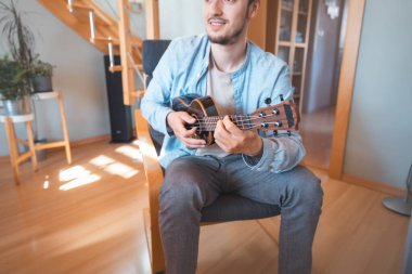 Cheerful man sits in a bright room, strumming a black ukulele. His light blue shirt and casual posture reflect a relaxed and joyful musical moment in a cozy setting. clipart