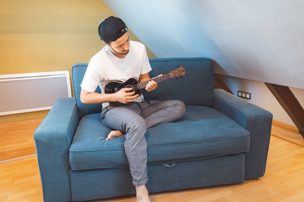 Man with talent sits on a blue sofa, carefully tuning a black ukulele. Musician is enjoying the feeling of playing the ukulele and immersing oneself in the creative spirit.