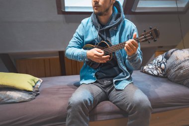 Musician with smile in a denim jacket and hoodie sits on a cozy bed, strumming a black ukulele. Enjoying the feeling of playing the ukulele and immersing oneself in the creative spirit. clipart