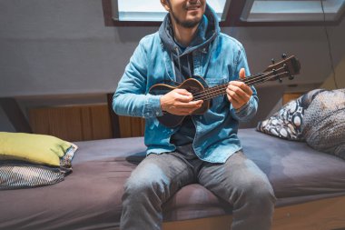 Kot ceketli ve kapüşonlu bir gülümseme giyen müzisyen rahat bir yatakta oturup siyah bir ukulele tıngırdatıyor. Ukulele çalmanın ve kendini yaratıcı ruha daldırmanın tadını çıkarıyorum..