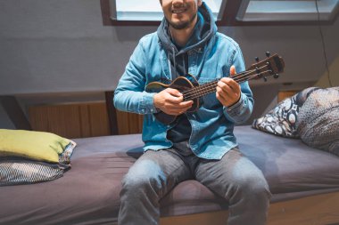 Kot ceketli ve kapüşonlu bir gülümseme giyen müzisyen rahat bir yatakta oturup siyah bir ukulele tıngırdatıyor. Ukulele çalmanın ve kendini yaratıcı ruha daldırmanın tadını çıkarıyorum..