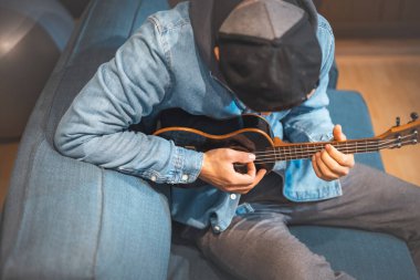 Musician in a denim jacket and hoodie sits comfortably on a blue sofa, playing a black ukulele. Boy with talent is  playing the ukulele in the safe environment of his home. Focus on music. clipart