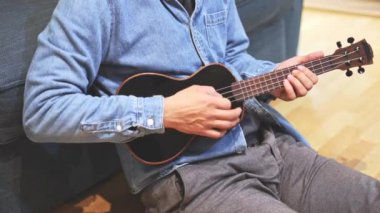 Gelişmekte olan bir müzisyenin siyah ukulele çalmayı öğrenip, müzik yeteneklerini derinleştirmesinin detaylı görüntüleri. Evlerinin konforunda telleri tıngırdatarak