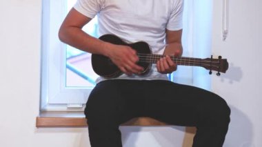 Gelişmekte olan bir müzisyenin siyah ukulele çalmayı öğrenip, müzik yeteneklerini derinleştirmesinin detaylı görüntüleri. Evlerinin konforunda telleri tıngırdatarak