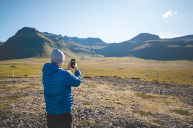 Traveler in a blue jacket and gray beanie captures a photo of the majestic volcanic mountains on the Snaefellsnes Peninsula in western Iceland. clipart