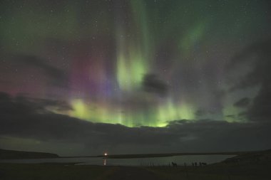 Batı İzlanda 'nın kırsal kesiminde çevreyi aydınlatan bir deniz feneriyle muhteşem bir Aurora Borealis. Kuzey ışıkları olarak İzlanda semalarında bir renk paleti yayıldı.