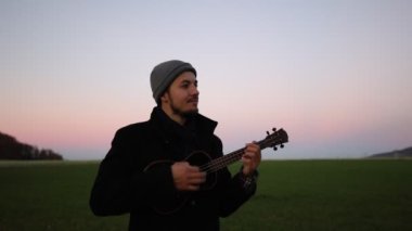 Tutkulu 26 yaşındaki müzisyen günbatımında gitar çalarak açık havada eğleniyor. Doğada olmak. Modern halk tarzında doğa ve müziğe duyulan sevginin bir karışımı. 4k video
