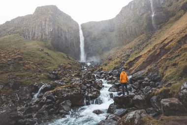 Turuncu ceketli gezgin, İzlanda, Snaefellsnes Yarımadası 'ndaki Grundarfoss şelalesinin önünde huşu içinde duruyor..