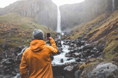 Turuncu ceketli bir gezgin Snafellsnes Yarımadası, İzlanda 'daki Grundarfoss şelalesinin güzelliğini yakalar. Şelalenin engebeli suları, yosunlu kayalıkları ve dağınık kayalıkları arasında durur..