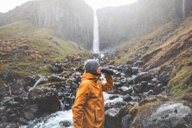 Turuncu ceketli gezgin, Snaefellsnes Yarımadası, İzlanda 'daki Grundarfoss şelalesinin yanında taze, kristal berrak buzul suyuyla dolu bir şişe içiyor..