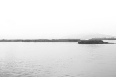 İzlanda 'da sakin suların, dağılmış kayalık adaların ve pürüzsüz bir şekilde gökyüzüne karışan sisli dağların yer aldığı siyah ve beyaz bir deniz manzarası yakalar..