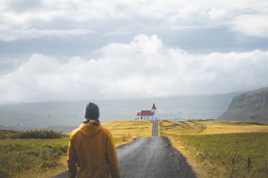 İzlanda 'nın Snaefellsnes yarımadasındaki Ingjaldsholskirkja Kilisesi' ne doğru sessiz bir yolda tek başına yürüyen bir yolcu. Yumuşak, bulutlu bir gökyüzünün altında altın tarlalarla çevrili.