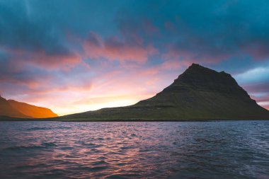 Güneş batarken İzlanda 'daki Snaefellsnes yarımadasında Kirkjufell Dağı. İkonik zirve, akşam gökyüzünün canlı renkleriyle çevrili sakin sulara yansıyor..