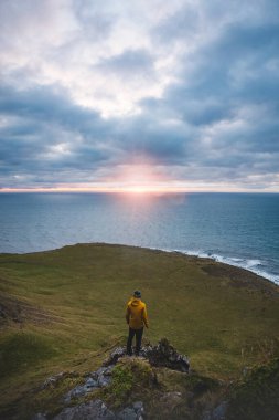 Sarı ceketli bir adam okyanusa bakan bir uçurumda duruyor. Güneş İzlanda 'nın batısındaki Snaefellsnes yarımadasında ufukta batıyor..