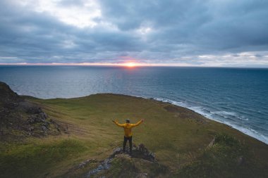 Sarı ceketli bir adam okyanusa bakan bir uçurumda duruyor. Güneş İzlanda 'nın batısındaki Snaefellsnes yarımadasında ufukta batıyor..