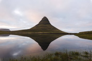 Kirkjufell, Batı İzlanda 'daki Snafellsnes Yarımadası' ndaki ikonik dağ, sakin sularda yansımasıyla görkemli bir şekilde duruyor, yumuşak, pastel bir gökyüzünün altında..