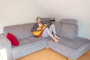 Çıplak ayaklı müzisyen gri bir kanepede uzanıyor, rahat bir atmosferde akustik gitar tıngırdatıyor, rahatlatıcı melodilerde kayboluyor ve müziğe derin duygusal bağlar kuruyor..