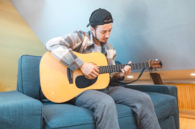Rahat tavırlı genç bir adam rahat mavi bir koltukta çıplak ayakla oturur, akustik gitar tıngırdatır. Müziğiyle bağlantı kurarken ifadesi memnuniyet ve odaklanma gösteriyor. Sıradan ve yaratıcı bir hava..