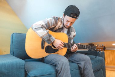 Rahat tavırlı genç bir adam rahat mavi bir koltukta çıplak ayakla oturur, akustik gitar tıngırdatır. Müziğiyle bağlantı kurarken ifadesi memnuniyet ve odaklanma gösteriyor. Sıradan ve yaratıcı bir hava..