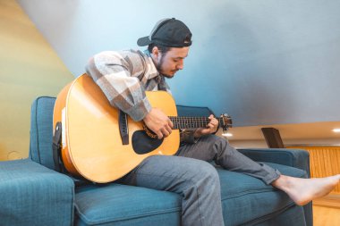 Rahat tavırlı genç bir adam rahat mavi bir koltukta çıplak ayakla oturur, akustik gitar tıngırdatır. Müziğiyle bağlantı kurarken ifadesi memnuniyet ve odaklanma gösteriyor. Sıradan ve yaratıcı bir hava..