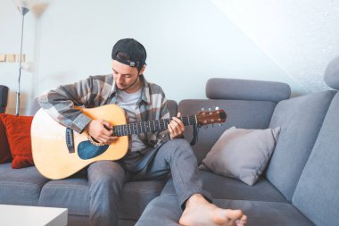 Rahat tavırlı genç bir adam rahat mavi bir koltukta çıplak ayakla oturur, akustik gitar tıngırdatır. Müziğiyle bağlantı kurarken ifadesi memnuniyet ve odaklanma gösteriyor. Sıradan ve yaratıcı bir hava..