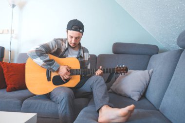 Rahat tavırlı genç bir adam rahat mavi bir koltukta çıplak ayakla oturur, akustik gitar tıngırdatır. Müziğiyle bağlantı kurarken ifadesi memnuniyet ve odaklanma gösteriyor. Sıradan ve yaratıcı bir hava..