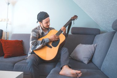 Rahat tavırlı genç bir adam rahat mavi bir koltukta çıplak ayakla oturur, akustik gitar tıngırdatır. Müziğiyle bağlantı kurarken ifadesi memnuniyet ve odaklanma gösteriyor. Sıradan ve yaratıcı bir hava..
