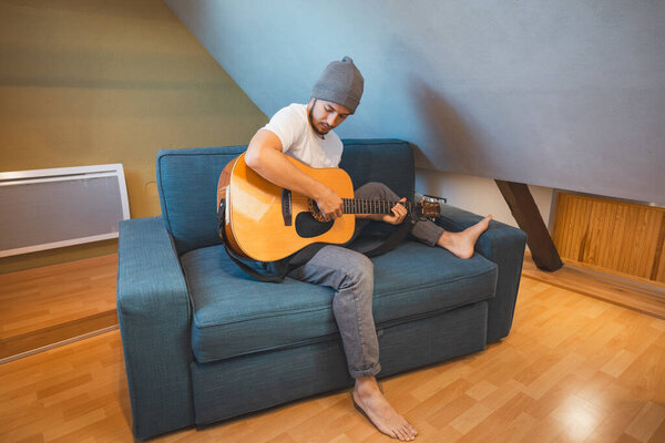 Musician 26 years old sits barefoot on a cozy blue couch in an attic room, strumming an acoustic guitar, lost in the melodies, creating a relaxed and intimate atmosphere filled with creativity.