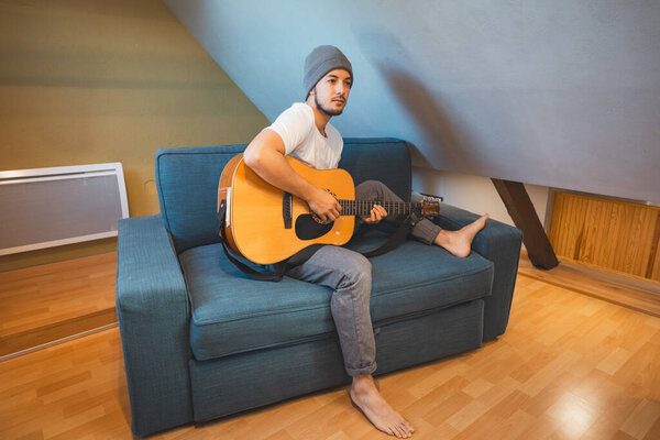 Musician 26 years old sits barefoot on a cozy blue couch in an attic room, strumming an acoustic guitar, lost in the melodies, creating a relaxed and intimate atmosphere filled with creativity.
