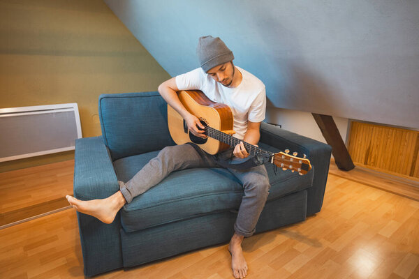 Musician 26 years old sits barefoot on a cozy blue couch in an attic room, strumming an acoustic guitar, lost in the melodies, creating a relaxed and intimate atmosphere filled with creativity.