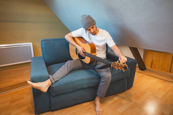 Musician 26 years old sits barefoot on a cozy blue couch in an attic room, strumming an acoustic guitar, lost in the melodies, creating a relaxed and intimate atmosphere filled with creativity.