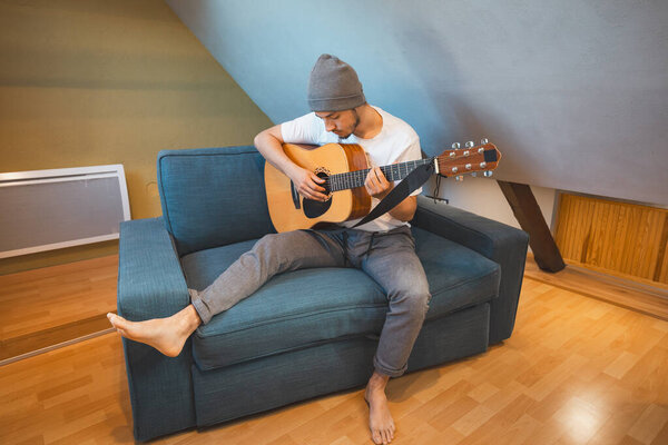 Musician 26 years old sits barefoot on a cozy blue couch in an attic room, strumming an acoustic guitar, lost in the melodies, creating a relaxed and intimate atmosphere filled with creativity.