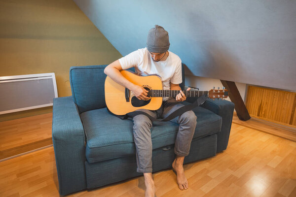 Musician 26 years old sits barefoot on a cozy blue couch in an attic room, strumming an acoustic guitar, lost in the melodies, creating a relaxed and intimate atmosphere filled with creativity.