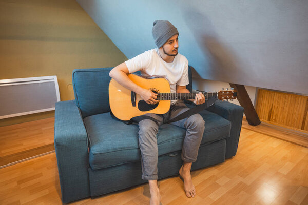 Musician 26 years old sits barefoot on a cozy blue couch in an attic room, strumming an acoustic guitar, lost in the melodies, creating a relaxed and intimate atmosphere filled with creativity.
