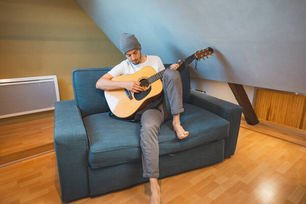 Musician 26 years old sits barefoot on a cozy blue couch in an attic room, strumming an acoustic guitar, lost in the melodies, creating a relaxed and intimate atmosphere filled with creativity.