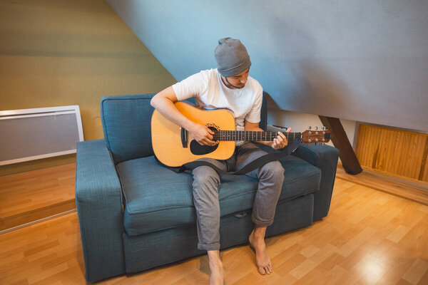 Musician 26 years old sits barefoot on a cozy blue couch in an attic room, strumming an acoustic guitar, lost in the melodies, creating a relaxed and intimate atmosphere filled with creativity.