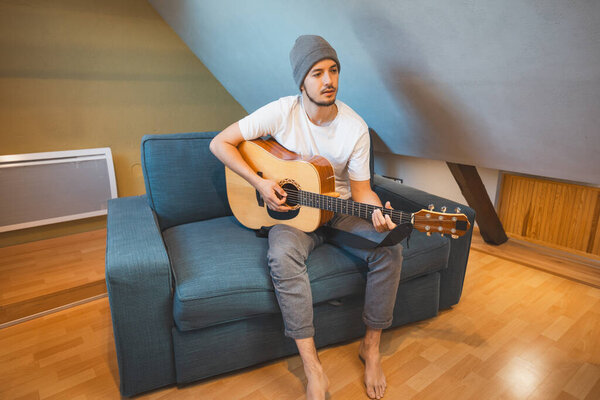 Musician 26 years old sits barefoot on a cozy blue couch in an attic room, strumming an acoustic guitar, lost in the melodies, creating a relaxed and intimate atmosphere filled with creativity.