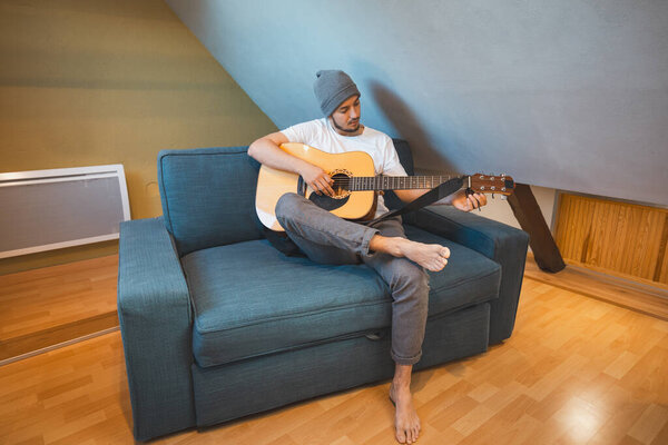 Musician 26 years old sits barefoot on a cozy blue couch in an attic room, strumming an acoustic guitar, lost in the melodies, creating a relaxed and intimate atmosphere filled with creativity.