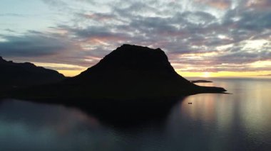 İzlanda 'nın batısındaki Grundarfjordur kasabası yakınlarındaki görkemli Kirkjufell dağının arkasındaki batan güneşin hava görüntüleri. Gece yarısı güneşinden gelen nefes kesici renkler. 4K video